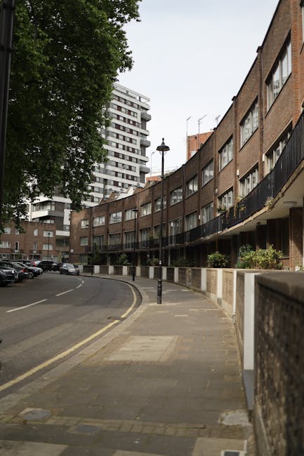 A low-angle view of a tall, multi-storey residential building with a dark brick facade, featuring multiple white-framed windows arranged in rows. Some of the windows have reflective glass showing the